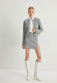 Harmaa tweed-takki ja hame-setti, jossa on sivupilkku, yhdistetty valkoisiin korkeisiin korkokenkiin. Takissa on kolme etupocketsia ja kiiltävät napit.