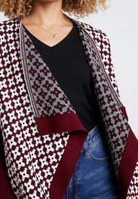 Burgunderfärgad och vit mönstrad kofta med stickad design, öppen front och en kontrasterande enfärgad kant längs kanterna. Bar över en svart topp.