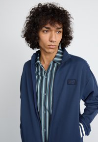 Navy zip-up jacket with white and blue striped accents, layered over a blue and white striped button-up shirt with a collar.