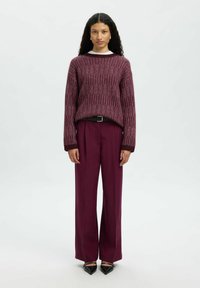 Femme avec de longs cheveux bouclés portant un pull texturé bordeaux, un pantalon large assorti, une ceinture noire et des chaussures à talons noirs, debout.