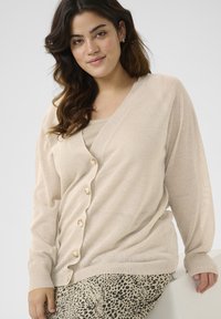 Beige lightweight cardigan with a deep V-neck, button front, and ribbed cuffs, paired with a textured tank top and patterned bottom.