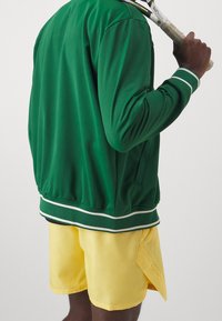 Groene sportjack met witte strepen aan de manchetten en zoom, gecombineerd met gele shorts met een zijsplit. De stof lijkt glad en ademend.