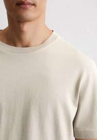 Beige Baumwoll-T-Shirt mit rundem Halsausschnitt und kurzen Ärmeln. Weiche Textur mit sichtbaren Nähten. Nahaufnahme, die sich auf den oberen Torso konzentriert.