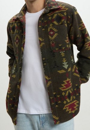 Mężczyzna ubrany w rozpiętą ciemnozieloną kurtkę z czerwonymi i żółtymi wzorami geometrycznymi, pod którą ma prostą białą koszulkę i jasnoniebieskie dżinsy.