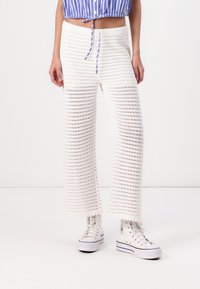 Weiß, weit geschnittene Hosen mit Häkelmuster und gestrickter Textur. Kombiniert mit weißen High-Top-Sneakern. Lockere Passform mit elastischem Bund.