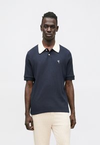 Laivastonsininen poolopaita, jossa on kerma valkoinen kaulus, lyhyet hihat ja pienellä kirjotulla sydänkuviolla. Valmistettu teksturoidusta kankaasta.