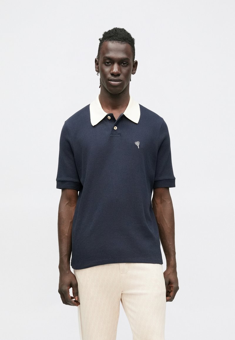 Laivastonsininen poolopaita, jossa on kerma valkoinen kaulus, lyhyet hihat ja pienellä kirjotulla sydänkuviolla. Valmistettu teksturoidusta kankaasta.