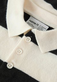 Polo majica s črnimi in kremnimi črtami, mehak material, zložen ovratnik in štirimi belimi gumbi, ki zagotavljajo zapenjanje. Vidna oznaka blagovne znamke.