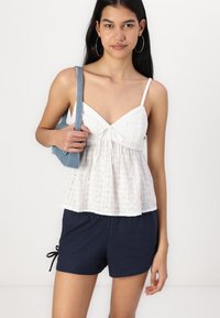 Balta, akis tinklui, maikutė su kaspinėlio detale, derinama su tamsiai mėlynais šortais, laikomais viena ranka. Modelis nešioja auskarus dideliais žiedo formos ir džinsinę petnešą.