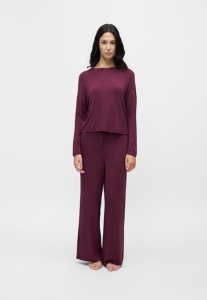 Femme debout pieds nus, portant un haut à manches longues assorti et un pantalon large de couleur violet foncé sur un fond clair uni.