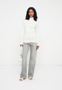 Donna che indossa un maglione bianco a collo alto con logo decorato, jeans grigi chiari a gamba larga, tacchi bianchi e tiene in mano una borsa bianca.