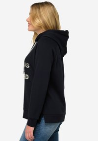 Sudadera navy con capucha ajustable, que presenta texto impreso en la parte frontal. Tela suave, corte relajado y puños acanalados. Combinada con jeans de mezclilla.
