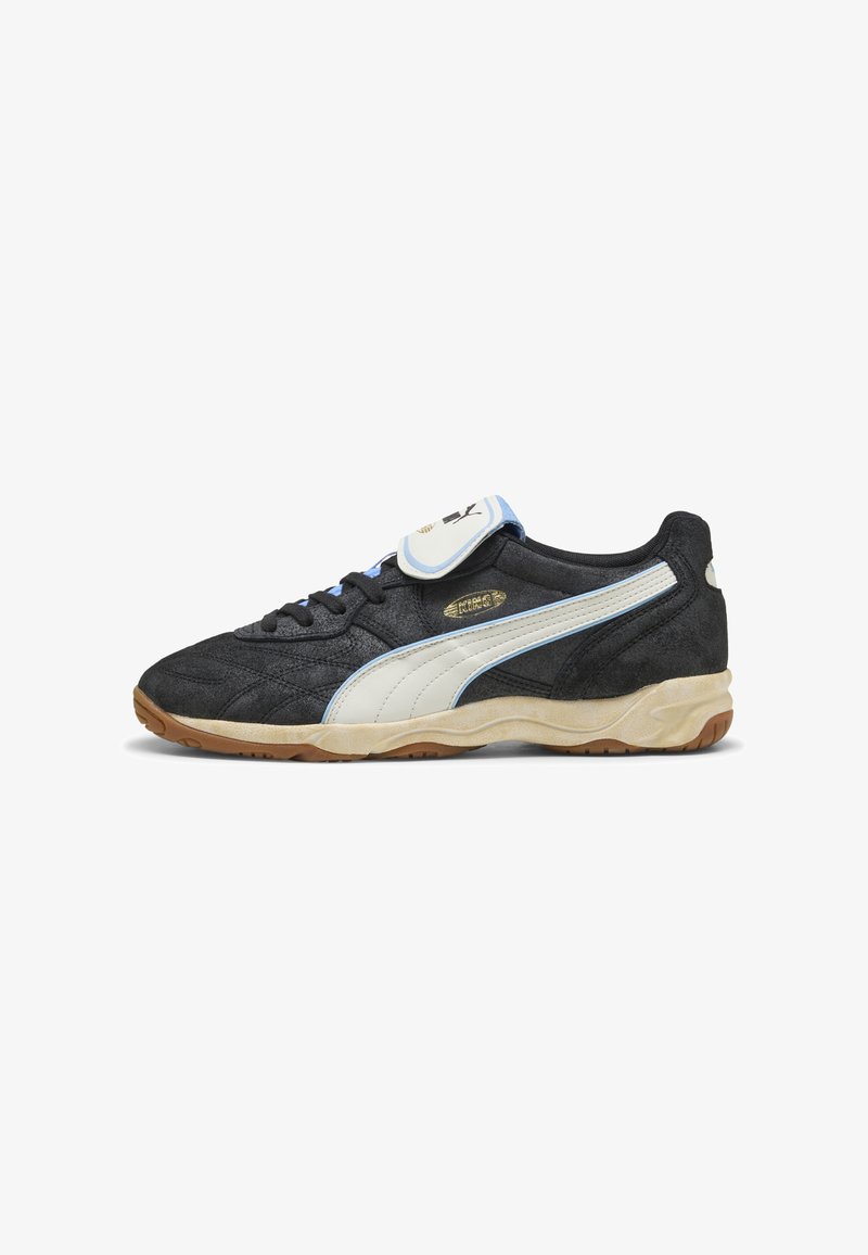 Sneaker Puma bianco e nero con suola in gomma, accenti azzurri chiari, lacci e un flap in velcro su sfondo bianco.