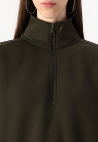 Dark green, high-neck top with a quarter-zip closure, made of soft, smooth fabric. Features minimalistic design and subtle stitching details.