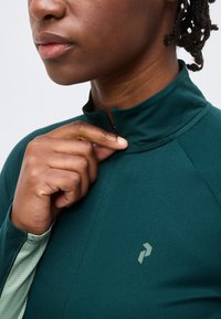 Άτομο με σκούρο δέρμα και πλεξούδες κουμπώνει ένα σκούρο πράσινο αθλητικό τοπ με ψηλό κολάρο και μικρό λογότυπο στο στήθος.