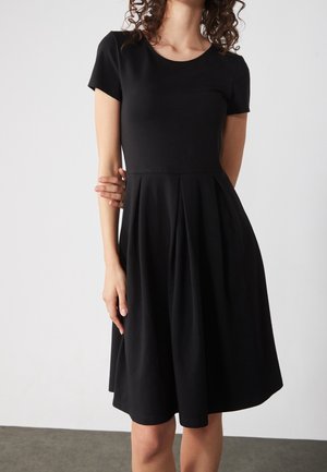 Frau trägt ein schwarzes knielanges Kleid mit kurzen Ärmeln, einem taillierten Oberteil und plissiertem Rock, steht vor einem weißen Hintergrund.