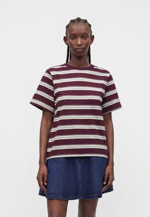 Femme aux cheveux tressés portant un t-shirt rayé bordeaux et blanc et une jupe en jean bleue, debout devant un fond clair uni.
