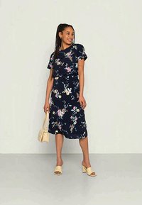 Vestido floral azul-marinho com mangas curtas, apresentando padrões de flores em rosa e verde. Combinado com sandálias bege e uma pequena bolsa bege.