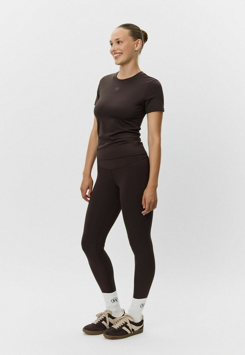 Brązowy zestaw odzieży sportowej z dopasowaną bluzką z krótkim rękawem i legginsami z wysokim stanem, w zestawieniu z brązowymi butami sportowymi i białymi skarpetkami do kostek.