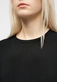 Γυναίκα με μακριά ξανθά μαλλιά που φοράει απλό μαύρο μπλουζάκι με στρογγυλή λαιμόκοψη, φωτογραφημένη από το πηγούνι έως το επάνω μέρος του στήθους με φόντο λευκό.