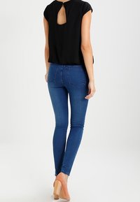 Černý top s výřezem na zádech, zkombinovaný s modrými skinny džínami. Džíny mají hladký denimový povrch a dvě zadní kapsy. Béžové vysoké podpatky.