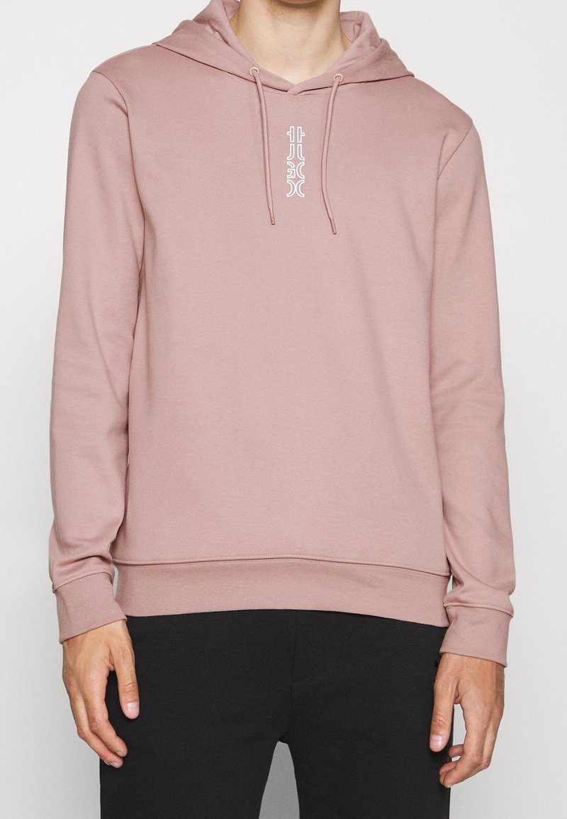Personne portant un sweat à capuche rose pâle avec des cordons de serrage à l'avant et un petit graphique blanc vertical sur la poitrine, associée à un pantalon noir.