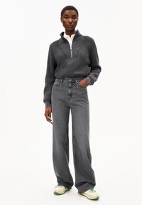 Grijze ribgebreide pullover met een halve ritskraag, gecombineerd met hoog-gesneden grijze wijde jeans en witte sneakers met groene accenten.