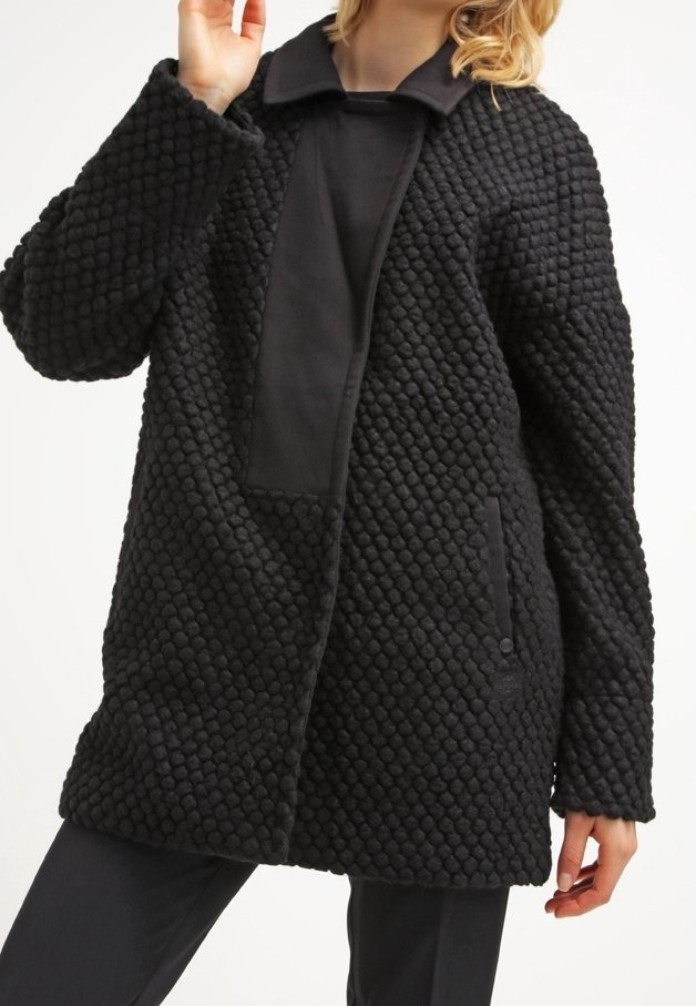 Abrigo negro texturizado con superficie acolchada, con un cuello con solapa, un bolsillo frontal y una silueta recta.