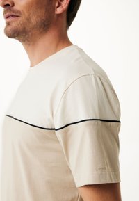 Béžové tričko s krátkým rukávem a kontrastním černým horizontálním pruhem přes hruď. Vyrobeno z hladkého bavlněného materiálu s uvolněným střihem.