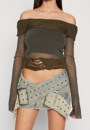 Donna che indossa un top in rete verde oliva consumato a spalle scoperte e una minigonna asimmetrica in denim chiaro con grandi cinture borchiate con occhielli.