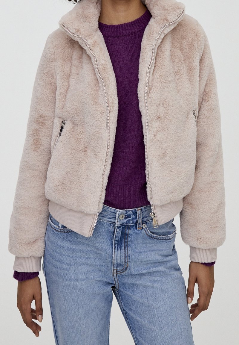 Faux fur bomberjakke i lyserød, med høj krave, frontlynlås og elastiske manchetter. Båret over en lilla sweater og jeans.