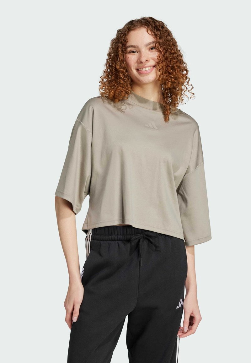 Camiseta corta de color gris claro, con mangas cortas y un escote alto. Presenta un logotipo sutil en la parte frontal. Combinada con pantalones negros.