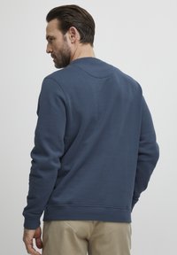 Mørkeblå hettegenser med rund hals, ribbestrikkede mansjetter og kant, samt en glatt tekstur, iført med beige bukser, sett bakfra.