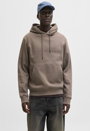 Hombre con sudadera beige de Jack & Jones, vaqueros grises y gorra azul, de pie frente a un fondo liso y claro.