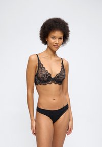 Γυναίκα με σγουρά μαλλιά που φοράει μαύρο δαντελένιο σουτιέν και ταιριαστό μαύρο εσώρουχο, στέκεται μπροστά σε απλό φωτεινό φόντο.