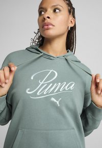Zelená mikina s kapucí, přední kapsou, stahovacími šňůrkami a velkým bílým logem Puma. Měkká tkanina a volný střih. Má pletený účes a náušnice.