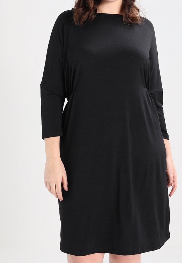 Femme portant une robe noire ample, longueur genoux, avec des manches trois-quarts, sur un fond clair uni.