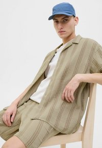 Ensemble de chemise à manches courtes et de shorts beige clair avec un tissu texturé et de subtiles rayures marron, associé à une casquette bleue. Modèle assis sur une chaise.
