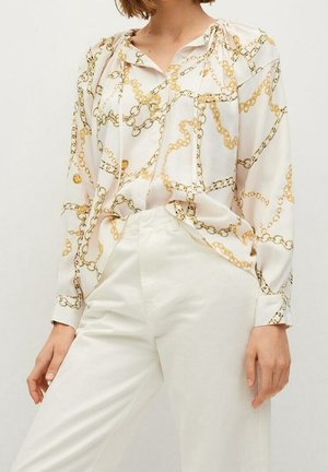 Kvinde iført hvid bluse med guld kædeprint og højtaljede hvide bukser, poserer mod en ensfarvet lys baggrund.