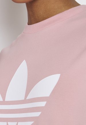 Detail osoby nosící světle růžové tričko s velkým bílým logem Adidas trefoil na hrudi.
