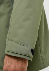 Olivengrüner Jackenärmel mit verstellbarem Klettverschluss, der teilweise ein tätowiertes Handgelenk zeigt.