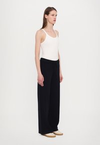 Hvit ribbet tanktopp kombinert med svarte, vide bukser med knyttet midje. Modell står mot en enkel hvit bakgrunn.