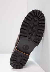 Semelle en caoutchouc noir avec des rainures profondes pour une traction, présentant un logo circulaire et un bord supérieur en cuir brun. Design texturé unique.