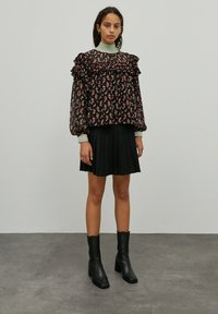 Zwart bloemetjesblouse met ruffles, doorschijnende stof en lange mouwen, gecombineerd met een geplooide zwarte rok en zwarte leren laarzen met hak.