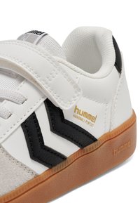 Hvid og sort Hummel-sneaker med velcrolukning og gummisål, viser logo og tekstureret indvendig krave.