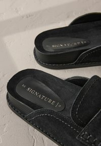 Sandales en suède noir au design plat, avec un logo embossé sur les semelles intérieures et des coutures contrastantes le long des bords.