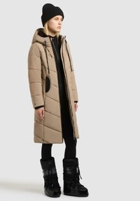 Beige vadderad lång kappa med huva, med svarta detaljer och dragkedja fram. Bärs med svarta leggings och höga svarta stövlar.