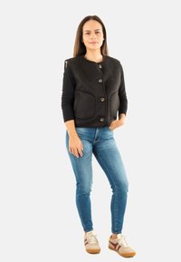 Gilet nero con bottoni, due tasche frontali, tessuto testurizzato, indossato sopra una camicia a maniche lunghe, abbinato a jeans blu e sneakers casual.