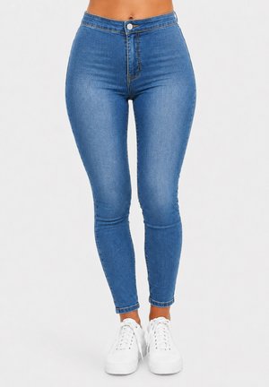 Vrouw draagt blauwe skinny jeans met hoge taille en witte sneakers, staand tegen een effen lichte achtergrond.