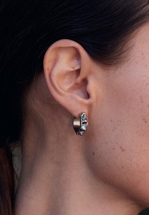 Silverfärgade ringörhängen med en texturerad design; har konformade accenter och en polerad finish, vilar på en modells öra.